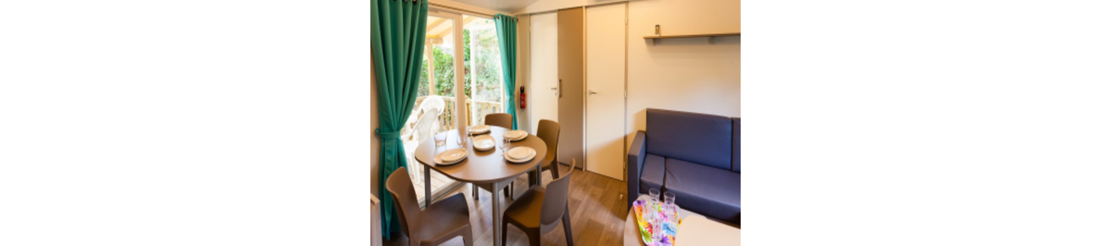 Compact dining and living space inside a holiday cabin is shown. A round table is set with plates and cups near glass doors that open to a small terrace. A blue sofa sits against the wall beside a shelf and closed interior doors. Bright turquoise curtains and warm wooden flooring create a welcoming, modern atmosphere.