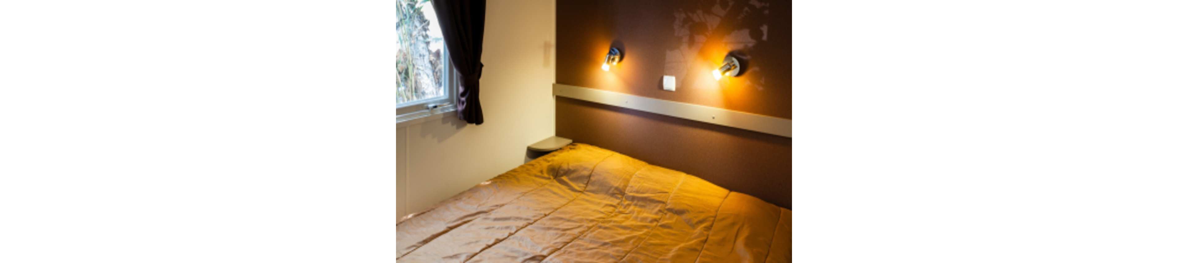 A neatly made bed with a tan or light brown comforter fills most of the small bedroom. Two wall-mounted reading lights cast a warm glow on the brown accent wall above the headboard. A window with dark curtains lets in some natural light from outside. The overall scene feels simple, tidy, and inviting, suitable for a hotel room or compact guest space.