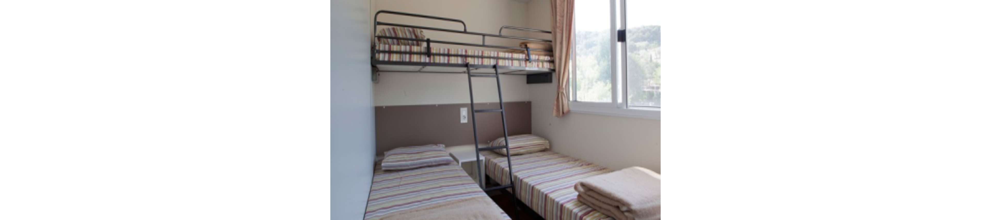The image shows a modest bedroom featuring a metal bunk bed above two single beds. All three beds have matching striped bedding and folded blankets placed neatly on top. A small bedside table sits between the lower beds, and a ladder leads up to the top bunk. Natural light enters through a large window with curtains, giving the room a bright and clean appearance.