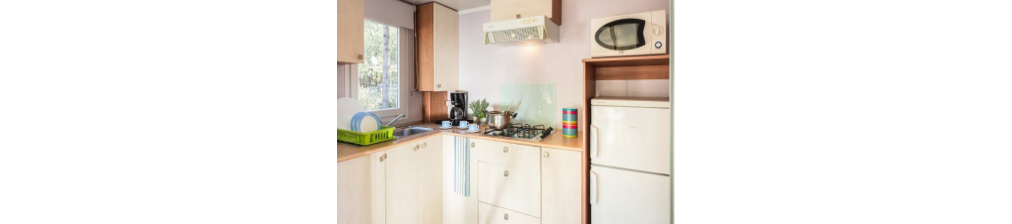 A compact modern kitchen features light wood cabinets and a gas cooktop with pots on the burners. A countertop holds a dish rack with plates, a coffee maker, and a small plant near a window. Above the stove is a range hood, while a microwave sits on a shelf above a white refrigerator. Soft pastel walls and simple decor create a clean, cozy atmosphere.