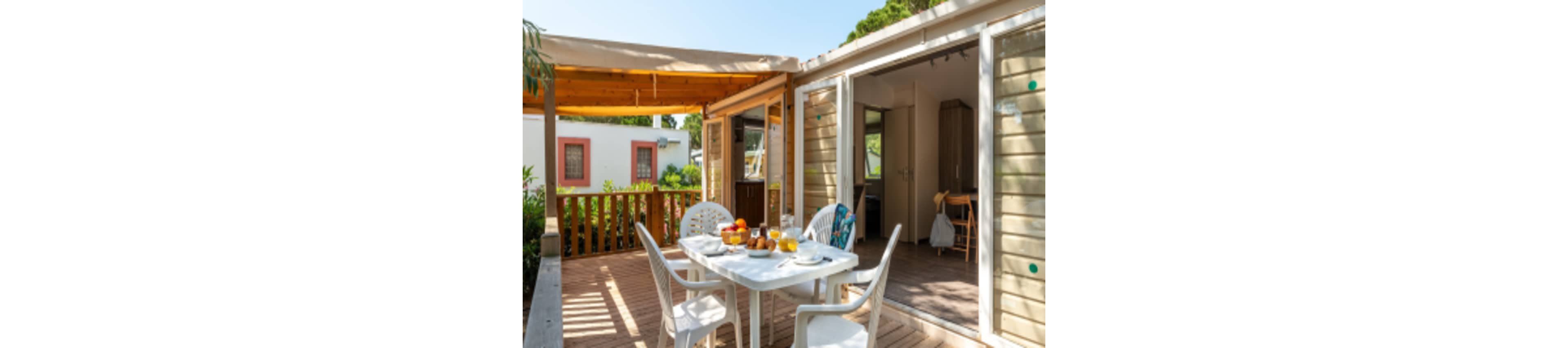 A cozy wooden terrace features a white plastic table set for breakfast with fruit, pastries, and drinks. The terrace is attached to a small cabin or bungalow with sliding glass doors leading to the interior. Sunlight filters through a partial wooden pergola roof, creating a warm and inviting atmosphere. Another small building is visible in the background, suggesting a holiday park or campsite setting surrounded by greenery.