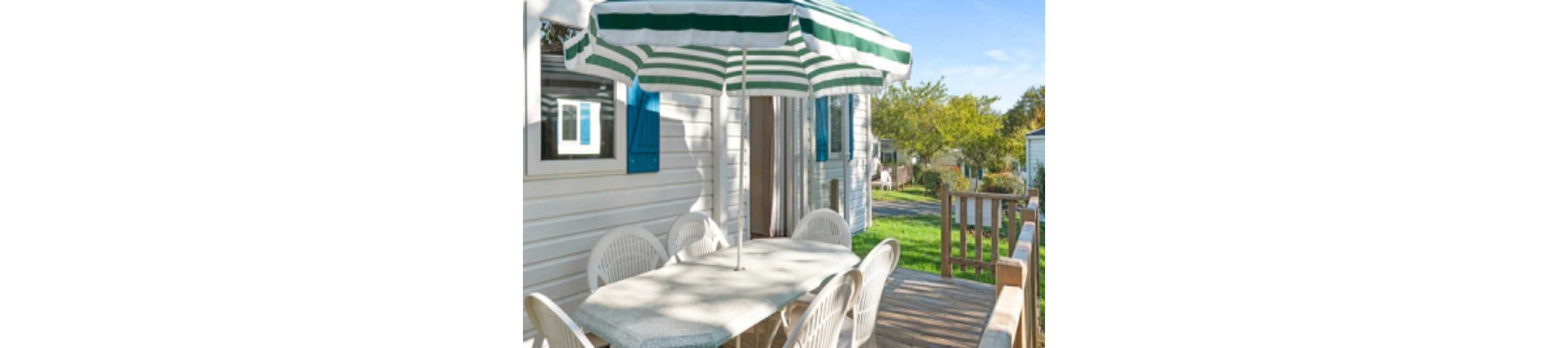 An outdoor dining area is set on a wooden terrace beside a white mobile home. A striped green and white patio umbrella shades a rectangular table surrounded by white plastic chairs. Blue shutters frame the nearby window, and the door to the mobile home stands open. Green grass, trees, and neighboring units are visible in the bright, sunny background.