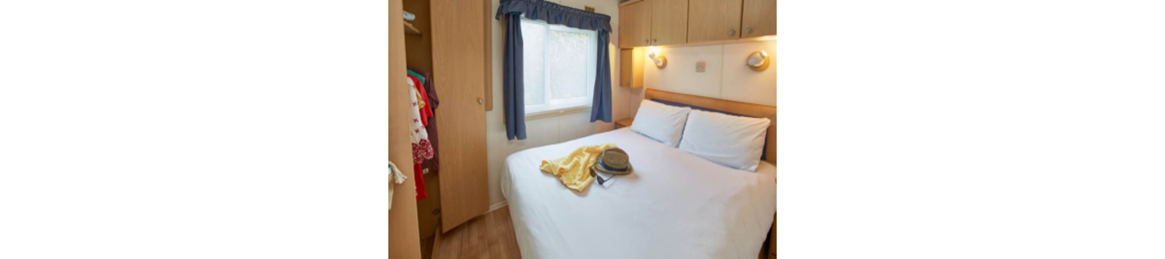 A compact bedroom features a neatly made double bed with white linens and two pillows. A hat and yellow garment are laid out on the bed, suggesting preparation for travel or a day out. Built-in wooden wardrobes and overhead cabinets provide storage around the bed. Natural light enters through a curtained window, while wall-mounted reading lights add a warm, inviting atmosphere.