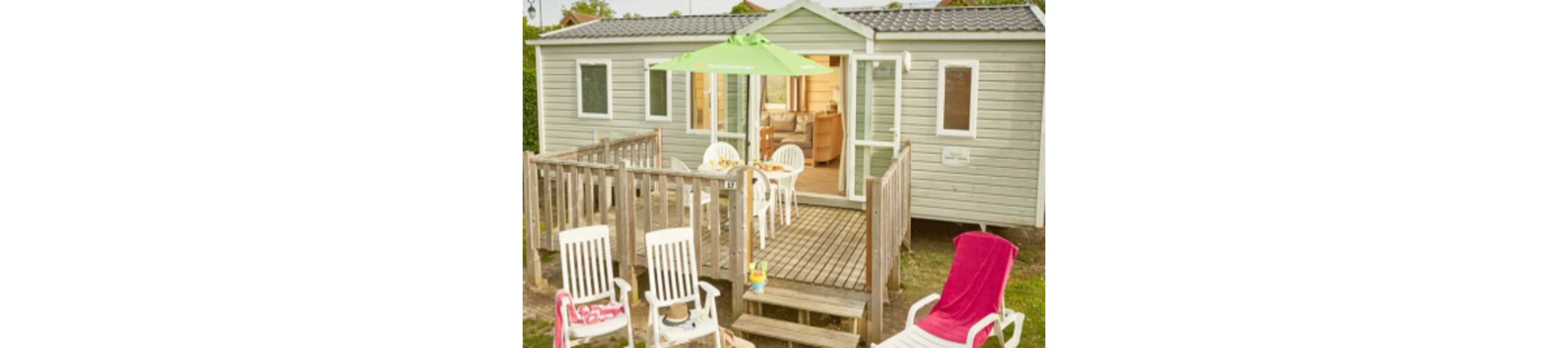 A light-colored mobile home features a wooden deck with outdoor seating and a green patio umbrella. White plastic chairs and a table set for a meal create a relaxed holiday atmosphere. A pink lounge chair sits on the grass in the foreground, suggesting a casual sunbathing spot. The open door and visible interior add a welcoming, family vacation feel.
