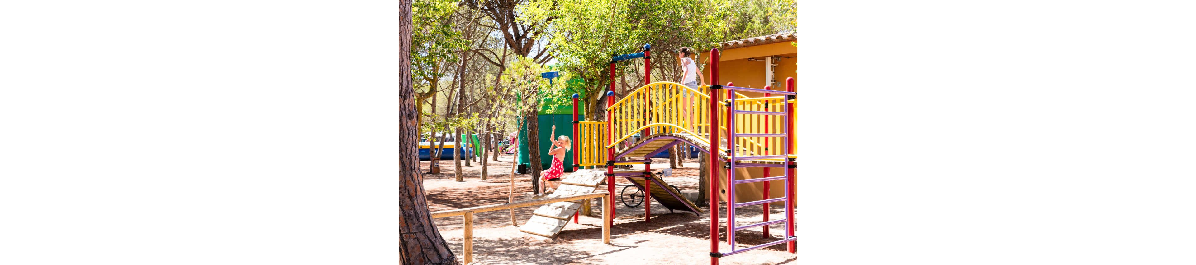 Two children play on a brightly colored playground structure in a wooded outdoor park. One child in a red dress swings from a rope while another stands on a yellow arched bridge. The play area is surrounded by tall trees, dappled sunlight, and nearby buildings painted in vivid colors. The scene feels lively and summery, emphasizing active play and childhood fun.