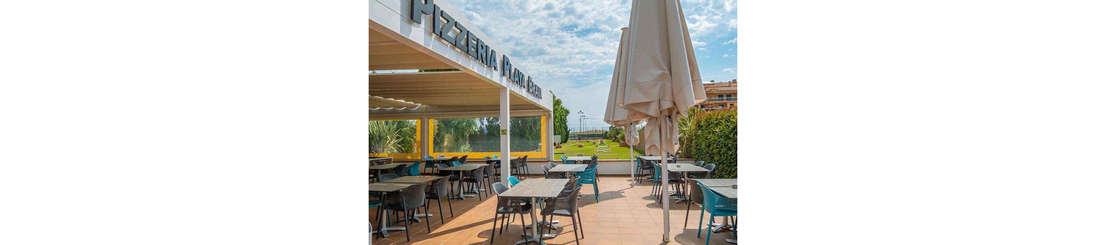 An open-air pizzeria terrace features neatly arranged tables and chairs under a large pergola. The sign reads "Pizzeria Playa Serena" above the covered seating area. In the foreground, folded patio umbrellas stand on a tiled floor, while the background reveals a manicured lawn, palm trees, and distant sea views. The scene is bright and sunny, suggesting a relaxed coastal dining atmosphere.