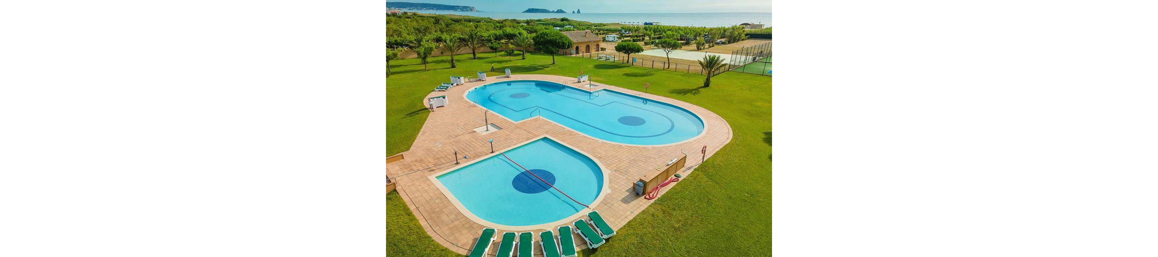 Aerial view of two connected outdoor swimming pools set in a large grassy resort area. Several green sun loungers line the poolside terrace, with clear blue water contrasting against the tiled deck. Palm trees, tennis courts, and low buildings stretch toward the distant coastline and sea. The scene suggests a tranquil holiday destination with ample recreational space and ocean views.