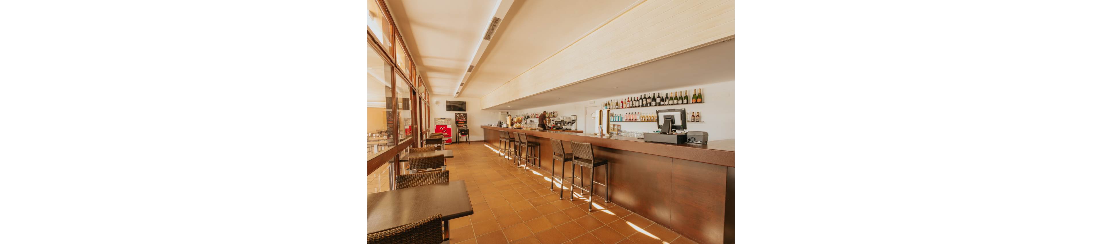 A spacious bar interior features a long wooden counter with several high stools neatly arranged. A bartender stands behind the bar, surrounded by shelves of bottles and glassware along the back wall. Large windows on the left flood the room with warm natural light, highlighting the tiled floor and minimalist furnishings. The overall atmosphere is clean, modern, and inviting, suitable for casual drinks or coffee.