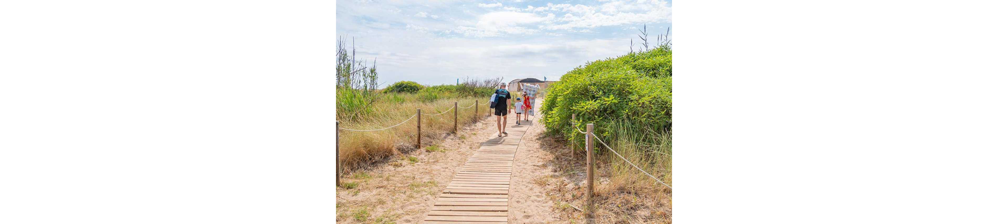 A family walks along a wooden boardwalk path toward the beach on a bright day. The scene is framed by sand dunes and tall grasses on either side of the walkway. The adults and children carry beach bags and towels, suggesting a relaxed seaside outing. The sky is lightly clouded, giving the image a warm, summery atmosphere.
