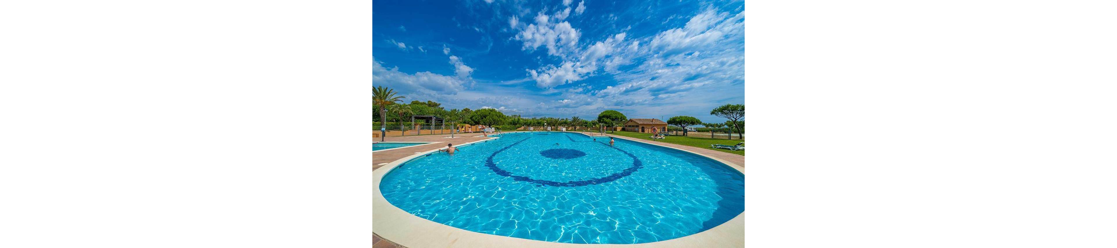 Large outdoor swimming pool with clear blue water is set in a sunny resort environment. A few people relax and swim in the spacious pool, surrounded by green lawns and palm trees. Sun loungers are arranged around the poolside, with small buildings and trees in the background. The bright sky is filled with scattered white clouds, creating a vibrant summer atmosphere.