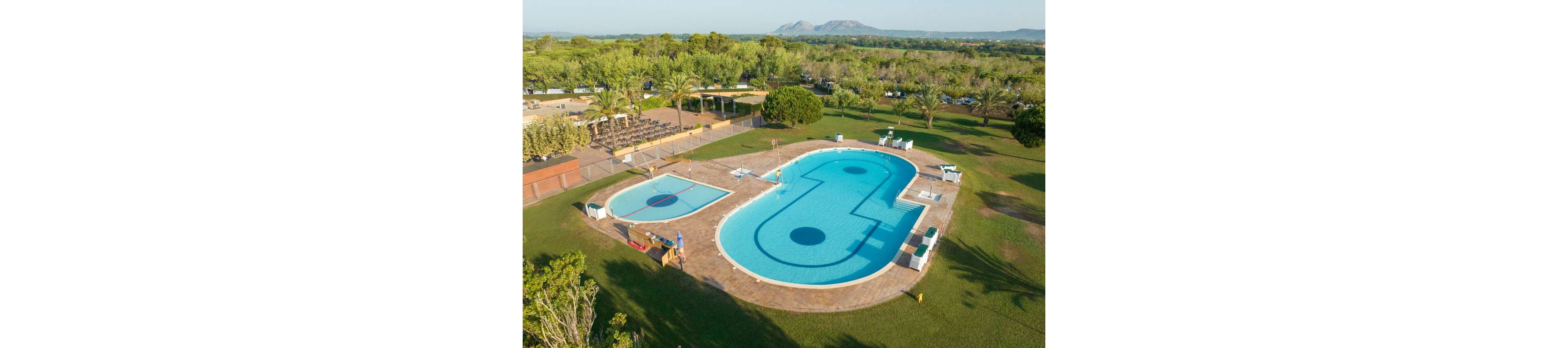 A large outdoor swimming pool with a unique curved shape is set in the middle of a spacious green lawn. Next to the main pool is a smaller shallow pool, likely for children, both surrounded by a tiled deck with sun loungers and umbrellas. A few people are visible near the pool area, and a shaded seating or dining area sits nearby under trees. In the distance, expansive fields and low mountains create a scenic natural backdrop under a clear sky.