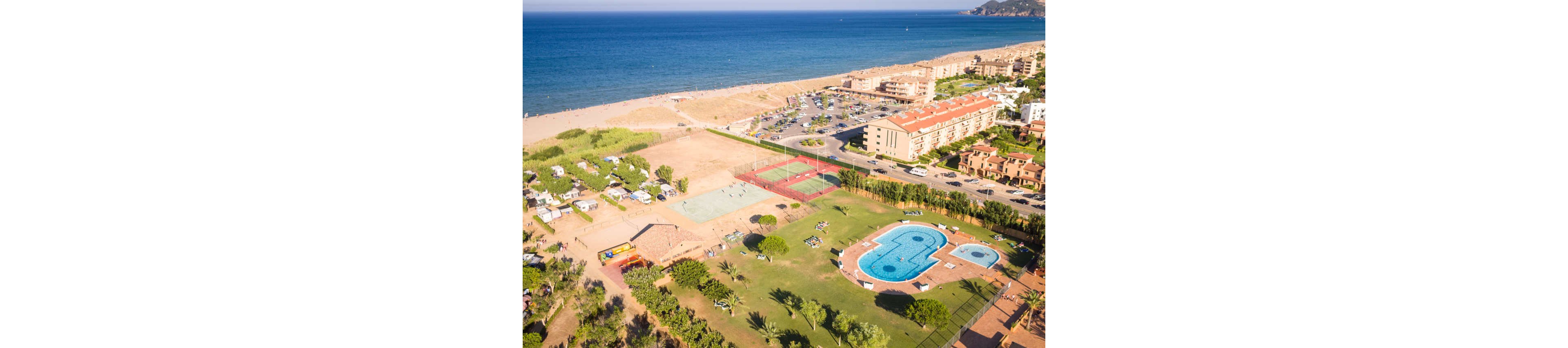A seaside resort complex is captured from above, showing a large outdoor swimming pool and landscaped green lawns. Nearby are sports courts, a sandy recreation area, and a campground with tents and caravans. Residential buildings and parked cars line the edge of the property, leading toward a wide sandy beach. The calm blue sea stretches into the distance under clear skies, creating a relaxed holiday atmosphere.