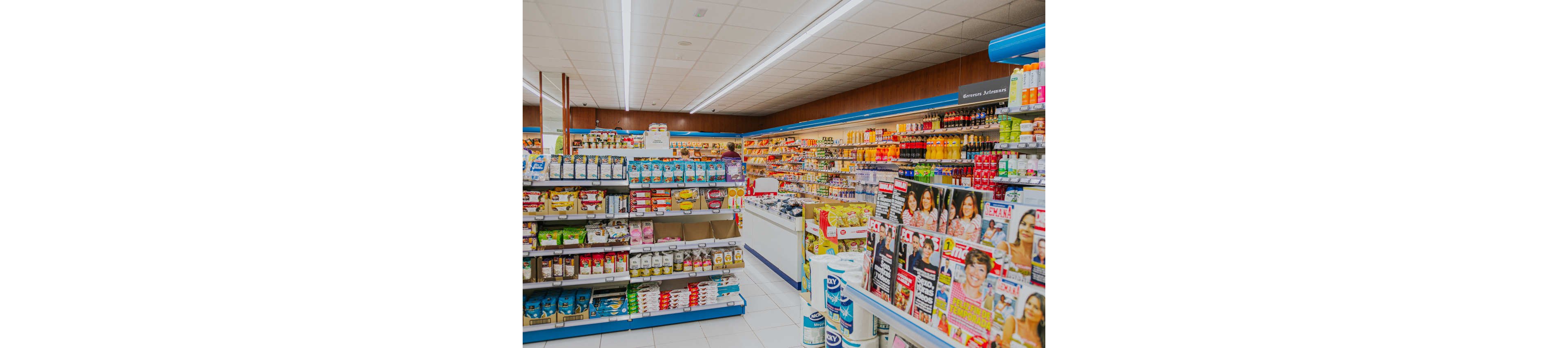 Wide view of a supermarket interior shows multiple aisles filled with groceries and household items. Shelves are stocked with boxed foods, canned goods, beverages, and paper products under bright fluorescent lighting. A magazine rack with colorful covers and portraits of people stands prominently in the foreground. A shopper is visible in the background, adding to the everyday retail atmosphere.