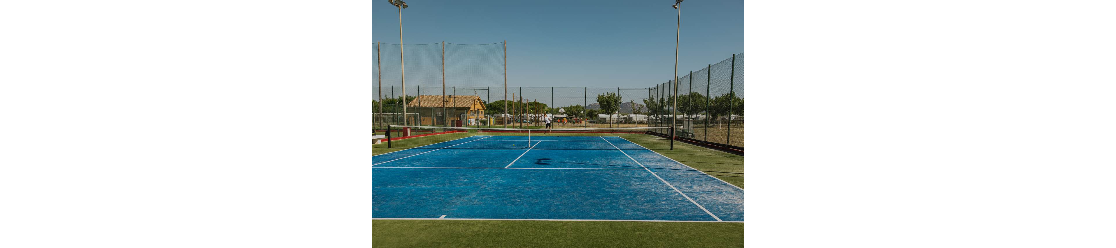 A bright blue tennis court sits empty under a clear daytime sky. The court is surrounded by tall chain-link fencing and multiple light poles. In the background, small buildings, trees, and parked vehicles are visible beyond the fence. The scene feels calm and quiet, suggesting an off-peak time at an outdoor sports facility.