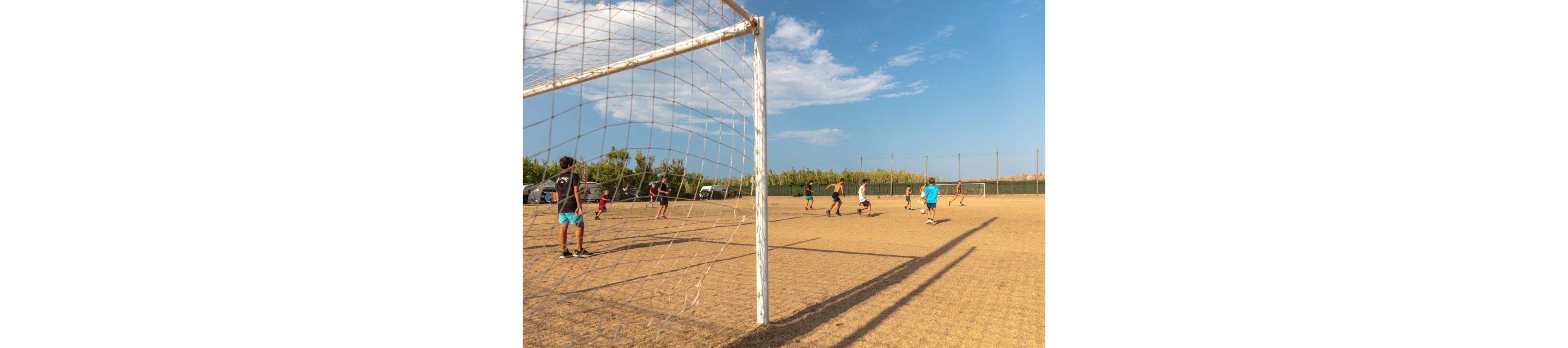 A group of teenagers play a casual soccer game on a dry, dusty field. The scene is viewed from behind the goal net, with players spread across the pitch chasing the ball. The sky is bright and mostly clear, suggesting a hot sunny day. Trees and fencing line the background, giving the impression of a local community or school sports ground.