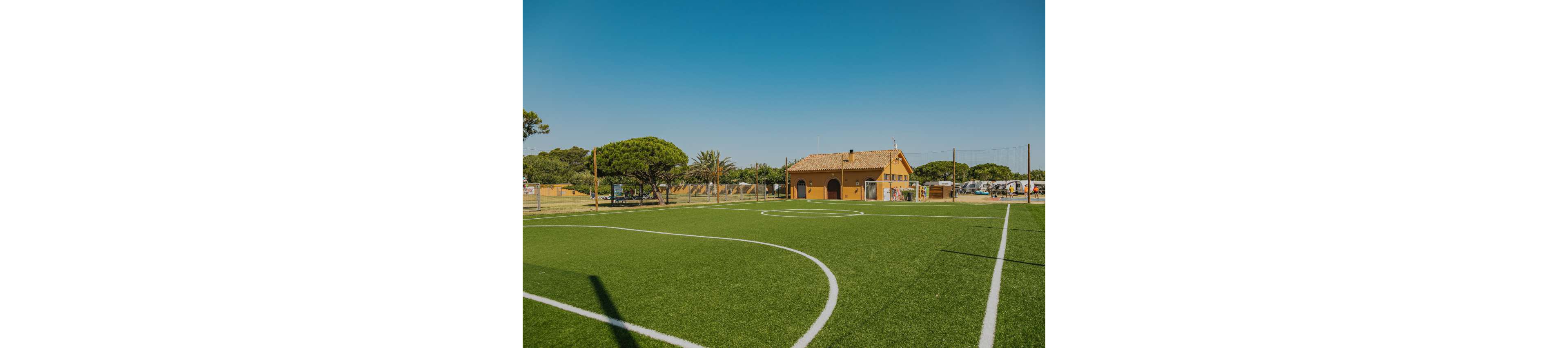 An empty artificial turf soccer field is shown under a clear blue sky. White field markings curve across the bright green surface, leading toward a small rustic building in the background. Trees and a few parked vehicles surround the area, suggesting a recreational or sports complex. The warm sunlight and open space create a calm, inviting atmosphere for outdoor activities.