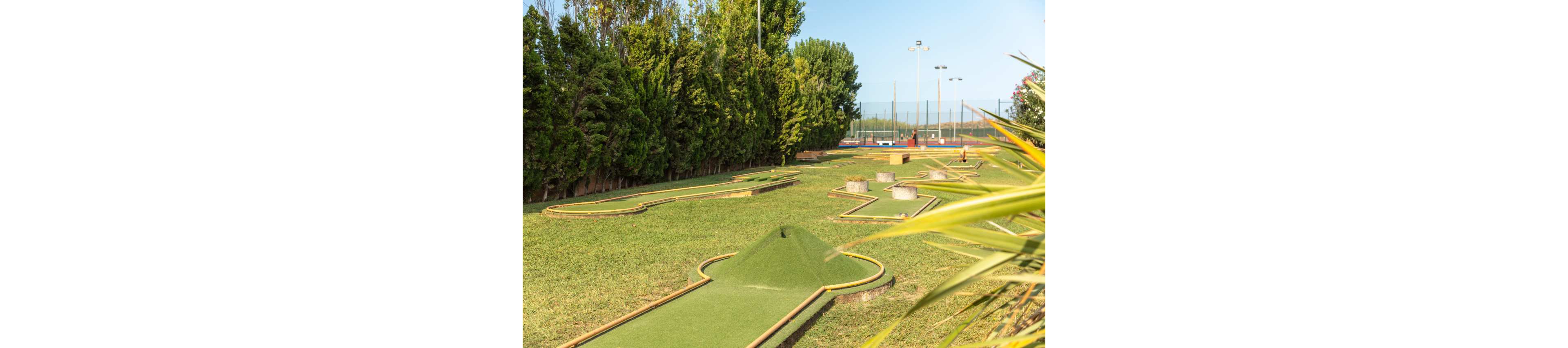 An outdoor mini golf course features several putting lanes laid out on a grassy field. Low wooden borders frame the artificial turf paths, with small hills and obstacles adding challenge. Tall green trees line one side, while sports courts and light poles are visible in the background. The bright daylight and clear sky create a relaxed, recreational atmosphere.