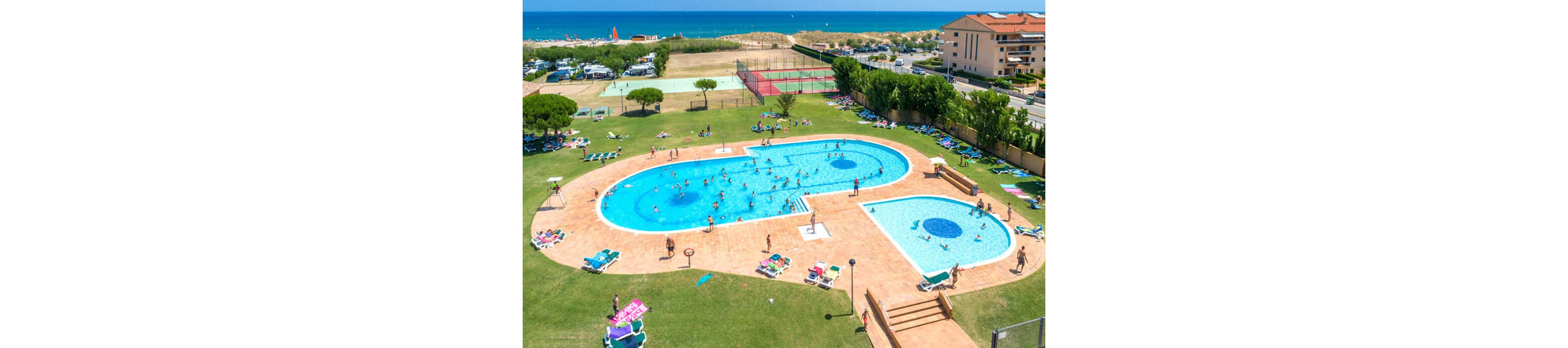 A large outdoor resort swimming pool is filled with people enjoying the water on a sunny day. Surrounding the pool are sun loungers with guests relaxing and sunbathing on the tiled deck. In the background, tennis courts, sandy dunes, and the blue sea are visible, along with nearby resort buildings. The bright daylight and clear sky create a vibrant, summery vacation atmosphere.