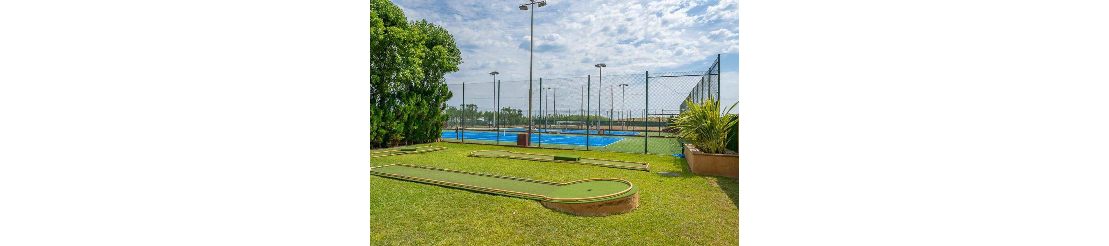 An outdoor recreation area features a small mini golf course in the foreground on a well-maintained grassy lawn. Behind it, several blue tennis courts are enclosed by tall chain-link fencing and lit by high light poles. Lush green trees and plants border the sports facilities, adding a natural backdrop. The scene is captured under a bright, partly cloudy sky, suggesting a pleasant day for outdoor activities.