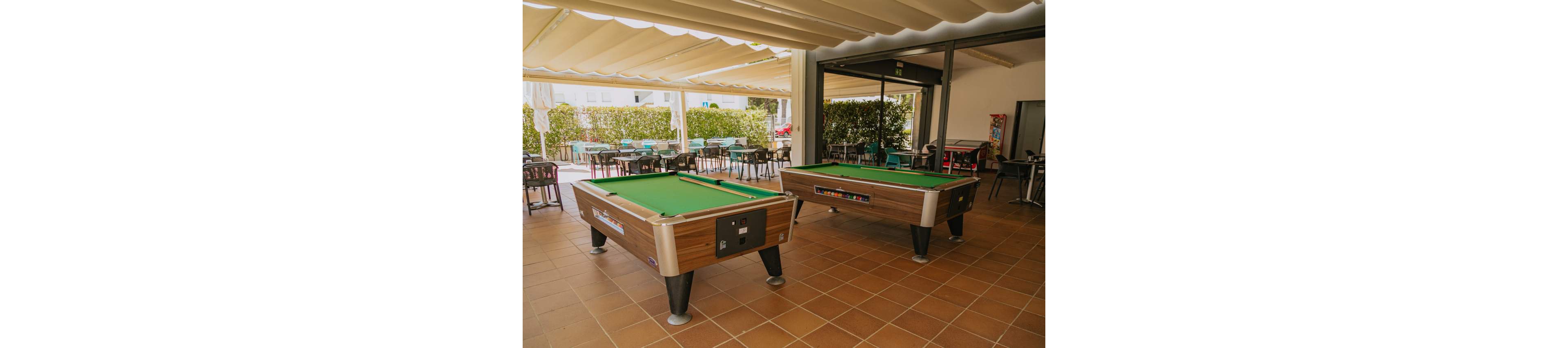 Two green-felt pool tables stand on a tiled patio under a covered pergola. In the background, tables and chairs are arranged for guests in an open-air seating area. Natural light filters through the canopy, creating a relaxed leisure atmosphere. The space appears ready for social gatherings and casual games.