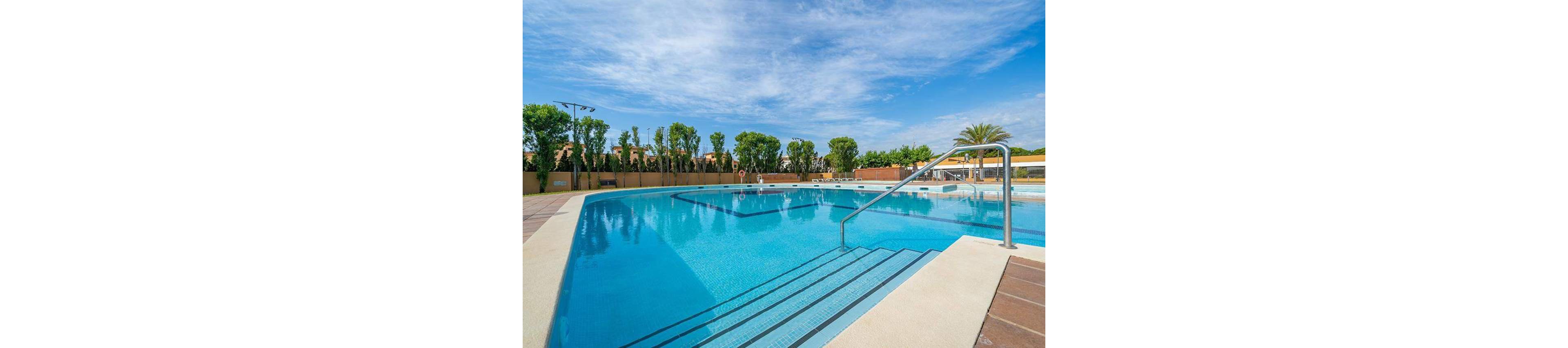A large outdoor swimming pool sits beneath a bright blue sky with scattered clouds. Wide entry steps and a metal handrail lead into the clear turquoise water. The pool is surrounded by tiled decking, safety buoys, and a fence lined with tall green trees. In the background, low buildings and palm trees suggest a resort or residential complex, creating a calm vacation atmosphere.