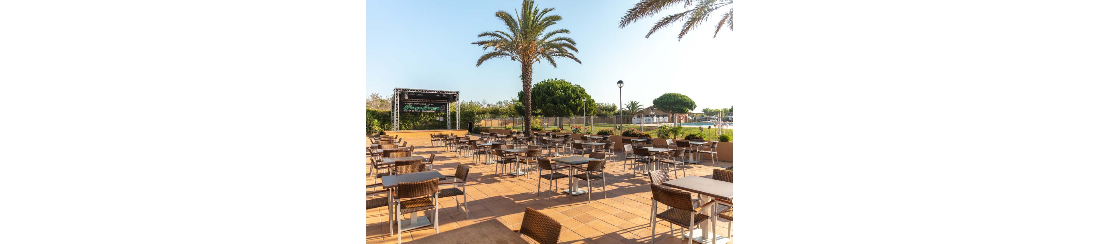 An empty outdoor dining terrace is arranged with rows of tables and chairs on a tiled floor. A small stage structure stands at the far left, suggesting a space for live performances or events. Tall palm trees and lush green landscaping frame the area under a clear blue sky. The scene feels calm and sunlit, ready to welcome guests for an open-air gathering.