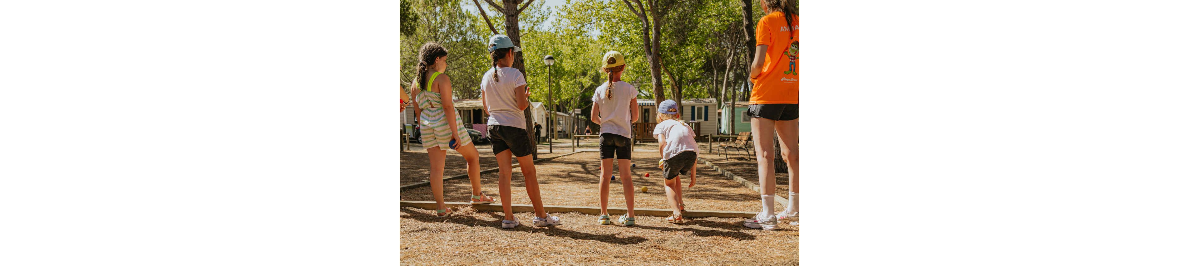 A group of children stand in a line playing an outdoor game on a dirt court. They appear to be in a shaded campsite or holiday park surrounded by trees and mobile homes. One child is in mid-throw while others watch and wait their turn. The scene is brightly lit with warm summer sunlight and casual clothing, creating a relaxed vacation atmosphere.