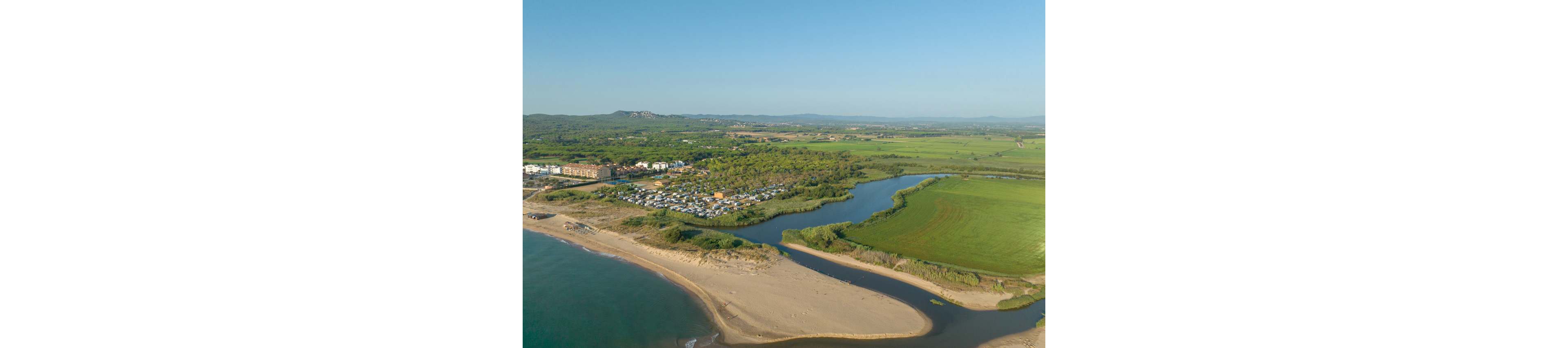 A wide aerial view shows a coastal campsite nestled between a sandy beach and green fields. A calm river or lagoon curves inland, separating the beach from the agricultural landscape. Numerous white vehicles and tents are visible among trees near the shoreline. In the distance, rolling hills and open countryside stretch under a clear blue sky.