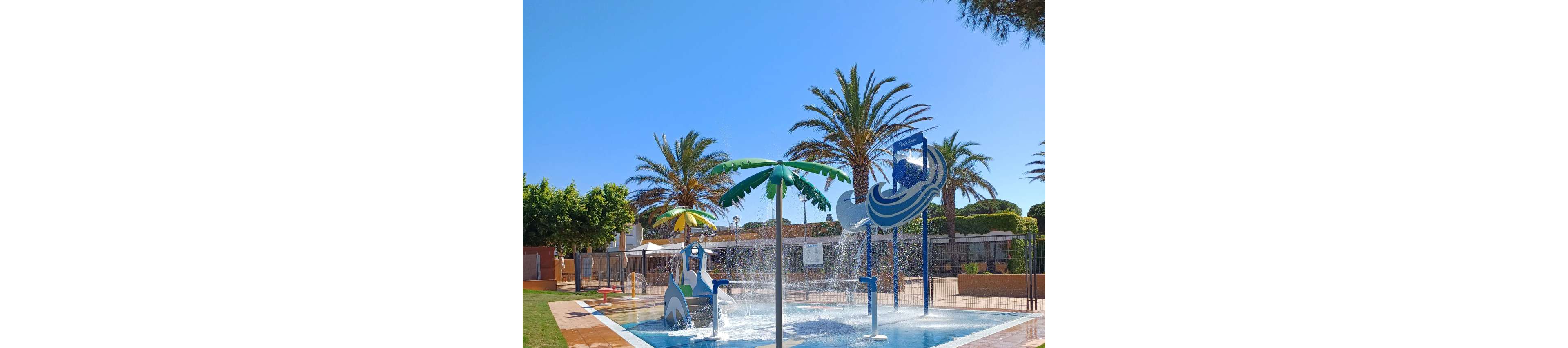 An outdoor splash pad features playful water structures shaped like palm trees and waves. Water sprays and fountains create an inviting scene under a clear blue sky. Tall palm trees and surrounding greenery frame the aquatic play area. Fences and nearby buildings suggest the splash park is part of a larger recreational facility.