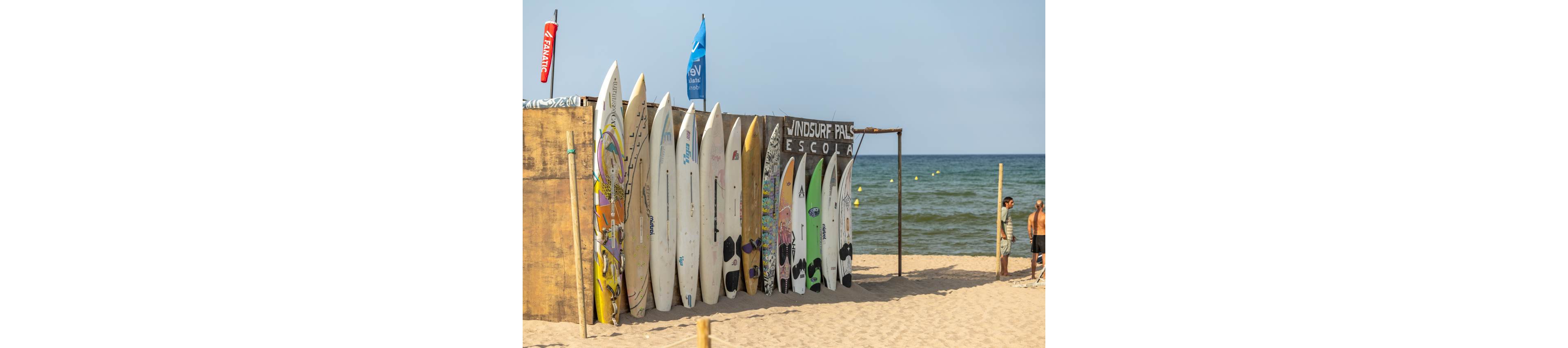 A row of colorful surfboards stands upright against a wooden wall on a sandy beach. A small surf school structure with flags and the word "ESCOLA" is visible beside the boards. Two men stand nearby talking while looking toward the sea. The calm ocean and clear sky create a relaxed summer atmosphere with a recreational, coastal vibe.