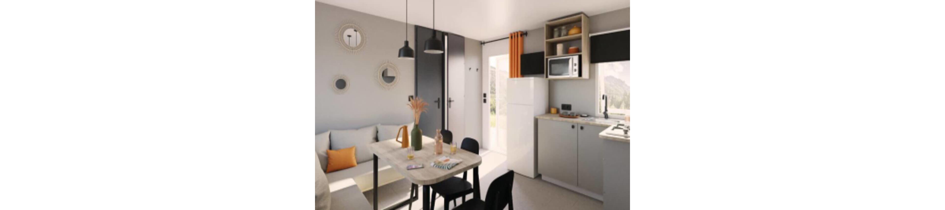 A small modern kitchen opens into a cozy dining and living nook. Light gray cabinets, a white refrigerator, and a built-in microwave define the compact cooking area. A wooden dining table with black chairs sits beside a cushioned bench with orange and gray pillows. Warm accents such as orange curtains, decorative vases, and round wall mirrors add contrast to the neutral interior.