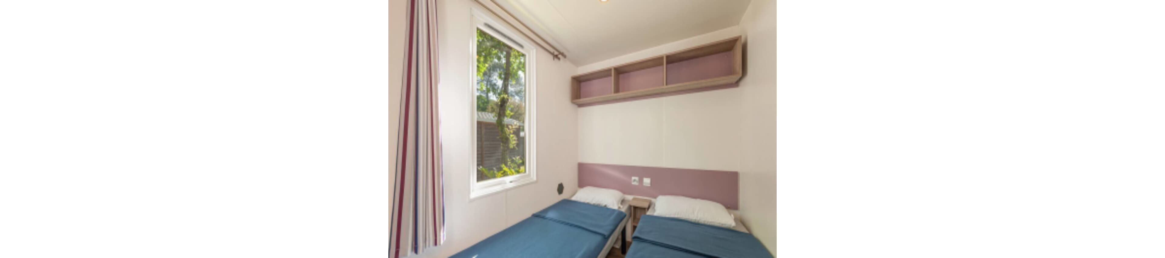 Compact bedroom featuring two single beds with blue bedding and white pillows. A large window lets in natural light and offers a view of greenery outside. Above the beds, open wooden shelves provide simple storage, and a small bedside table sits between the beds. The room has light-colored walls with a soft pink accent panel and striped curtains for a clean, modern look.