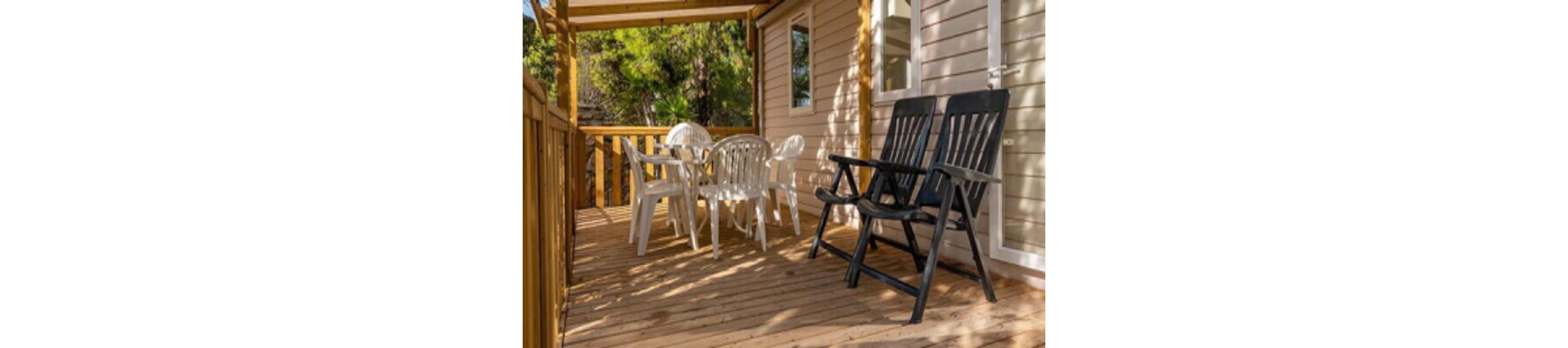 A cozy wooden terrace features a set of white plastic chairs around a round table and two black reclining chairs in the foreground. The deck is attached to a light-colored mobile or modular home with a window and door visible. Warm sunlight filters through the wooden railing and roof structure, creating inviting shadows on the floor. Green trees in the background suggest a peaceful, natural setting ideal for relaxing or outdoor dining.