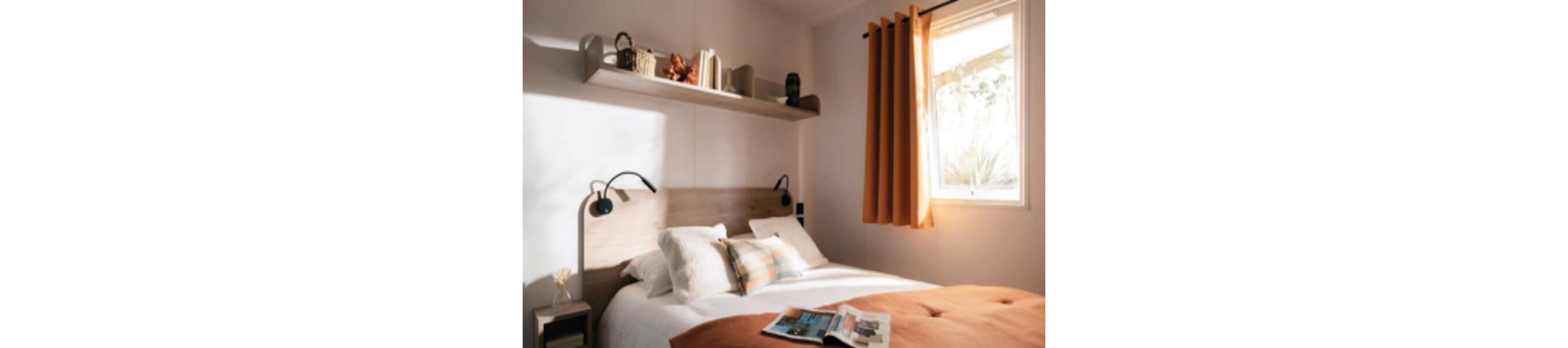 A neatly made double bed with white pillows and a plaid accent cushion sits against a light wooden headboard. Warm sunlight streams through a window with orange curtains, casting a soft glow across the room. A floating shelf above the bed holds books and decorative objects, adding a personal touch. A magazine lies open on the rust-colored duvet, enhancing the relaxed and inviting atmosphere.