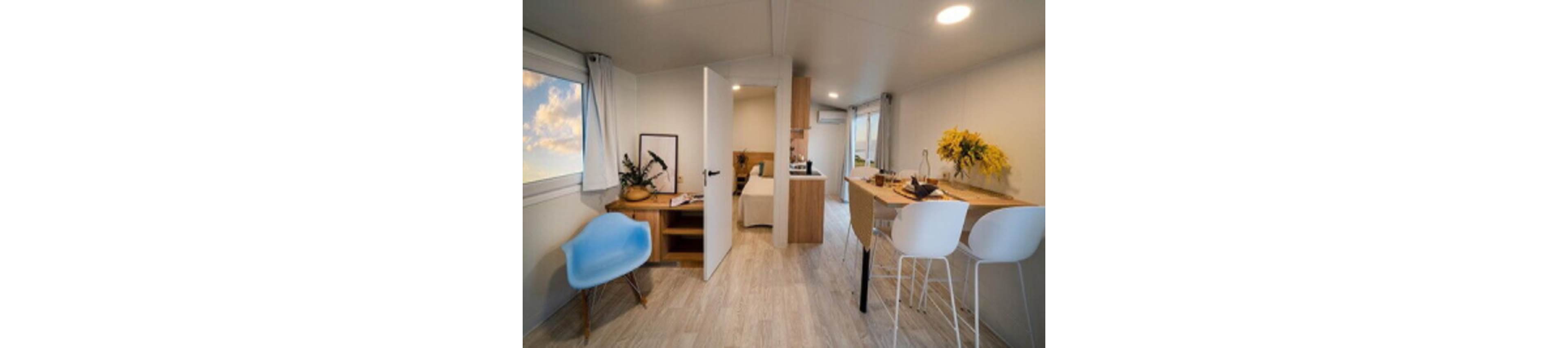 Bright modern tiny home interior featuring an open-plan layout. A compact kitchen and dining bar with white stools sit opposite a wooden countertop decorated with yellow flowers. In the foreground, a blue accent chair and small desk area add a cozy touch near a large window with sky views. An open door reveals a simple bedroom, emphasizing efficient use of space and minimalist design.