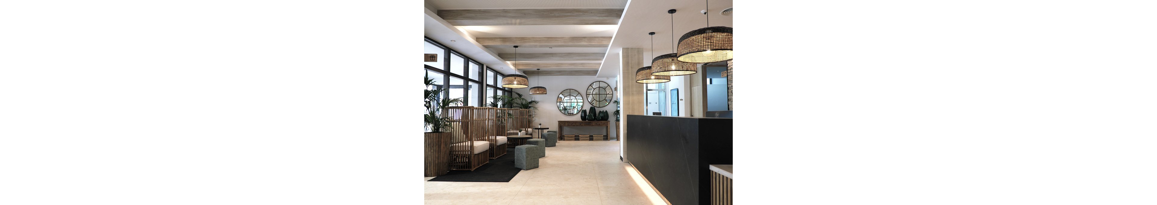 Contemporary hotel lobby interior featuring a sleek black reception desk and light tiled flooring. Wooden slat seating with white cushions lines one side, complemented by woven pendant lights overhead. Large round mirrors and decorative vases sit on a console table at the back wall, while potted plants add greenery throughout the space. Floor-to-ceiling windows bring in natural light, creating an airy and welcoming atmosphere.