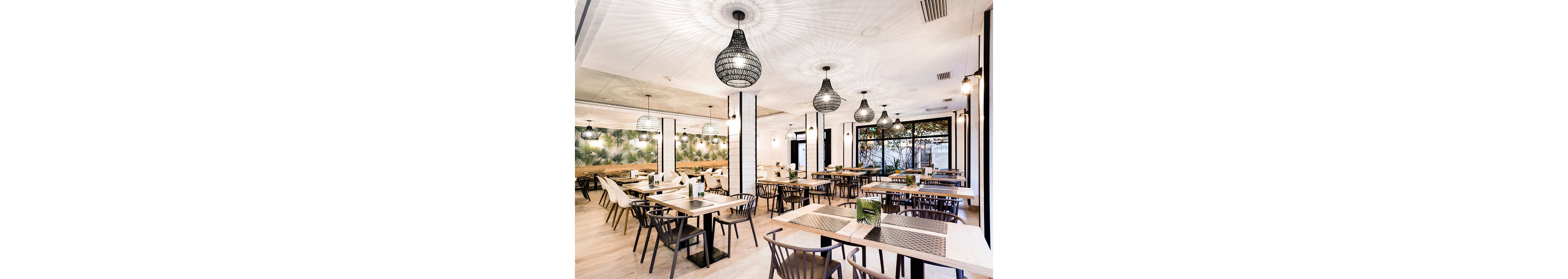 Spacious modern restaurant interior featuring neatly arranged wooden tables and chairs. The room is brightly lit with large woven pendant lights casting patterned shadows on the white ceiling. A long wall mirror and decorative greenery add depth and warmth to the space. Floor-to-ceiling windows bring in natural light and offer a view of trees outside, while table settings remain minimal and elegant.