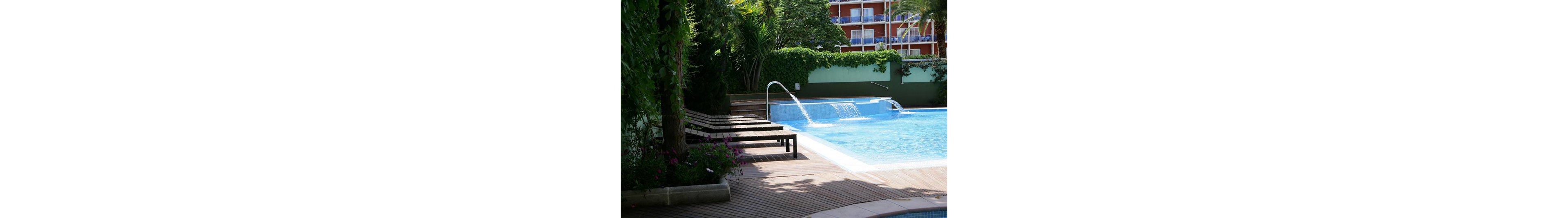 An inviting outdoor swimming pool is surrounded by wooden decking and several dark lounge chairs. Water cascades from metal spouts into the clear blue pool, creating a refreshing atmosphere. Lush green plants and ivy-covered walls frame the scene, with a multi-story hotel building visible in the background. The setting suggests a tranquil vacation spot on a bright, sunny day.