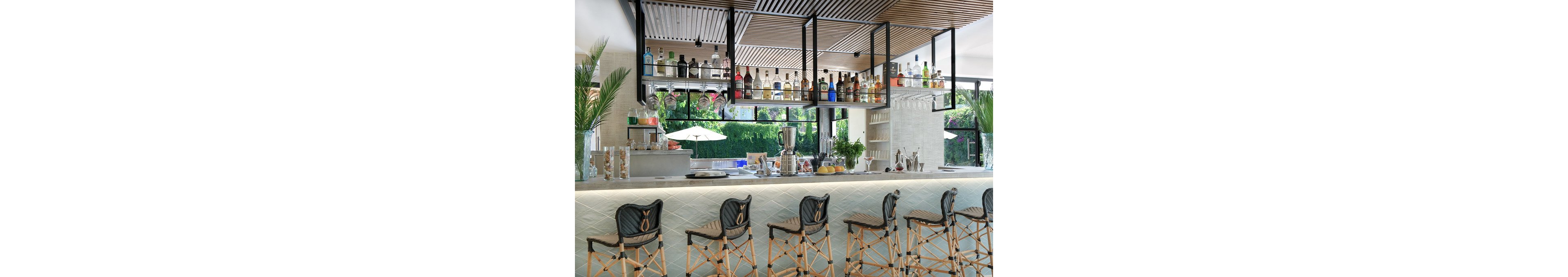 A contemporary bar counter is lined with woven high stools in a bright, airy setting. Bottles of liquor and spirits are neatly arranged on suspended shelves above the bar. Fresh ingredients, glassware, and bar tools are displayed on the countertop, suggesting cocktail preparation. Large windows reveal a lush garden with umbrellas and greenery, enhancing the relaxed, resort-like atmosphere. The overall design features light tones, natural textures, and clean architectural lines.