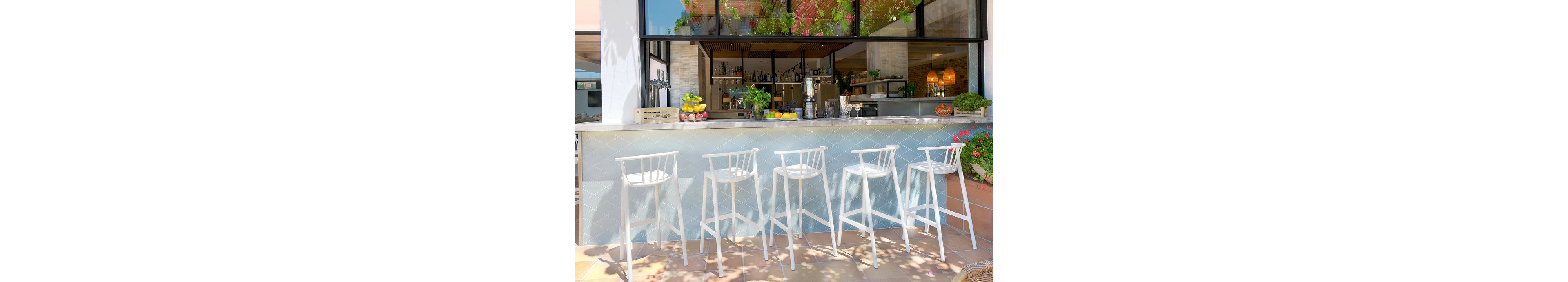 A bright outdoor bar features a row of five tall white stools lined up along a tiled counter. The bar area is open to the outside, with large windows revealing shelves of bottles and glassware inside. Fresh fruits and plants decorate the countertop, adding color and a relaxed, summery feel. Soft natural light and shadows from nearby foliage create an inviting atmosphere for guests.