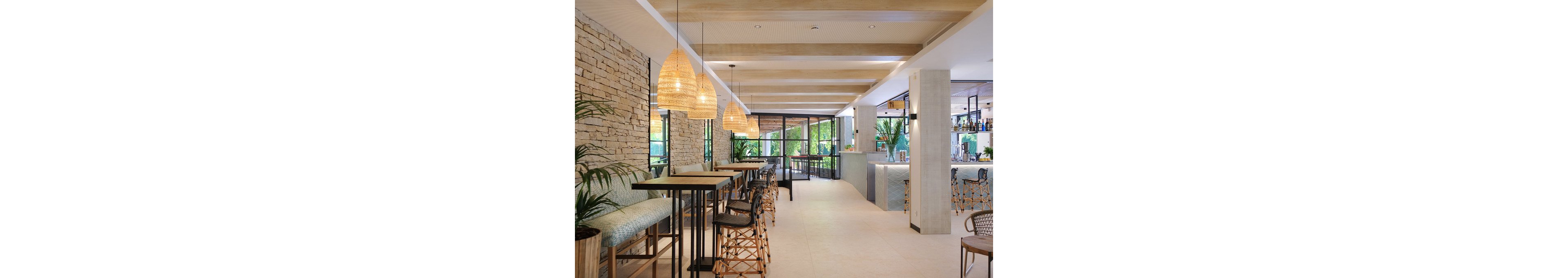Contemporary bar lounge interior featuring high tables, cushioned benches, and woven bar stools. Large wicker pendant lights hang from a beamed ceiling, casting warm light over the seating area. Stone accent walls, indoor plants, and floor-to-ceiling windows create an airy, tropical atmosphere. A sleek bar counter with bottles displayed in the background completes the relaxed, resort-style setting.