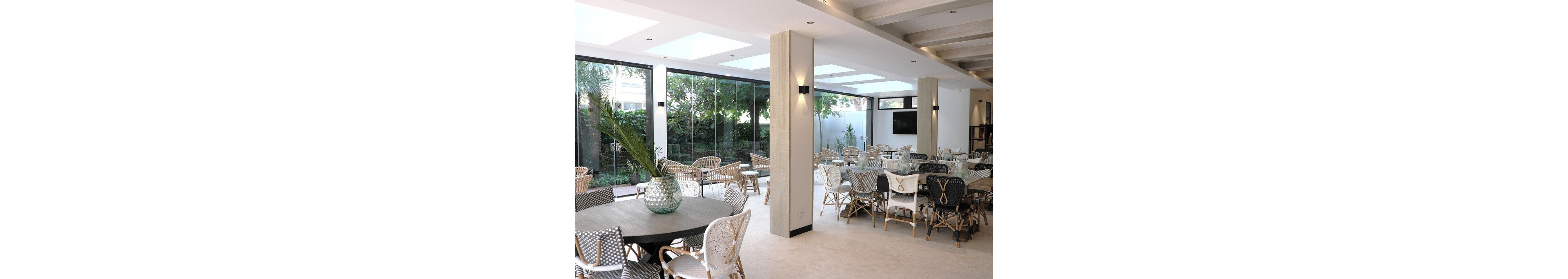 Spacious modern dining area features woven chairs and round tables arranged in an open-plan layout. Large floor-to-ceiling glass windows reveal lush greenery outside, bringing natural light into the room. Neutral tones, light wood accents, and minimalist decor create an airy, contemporary atmosphere. A decorative glass vase with foliage sits on a central table, adding a fresh, organic touch to the interior.