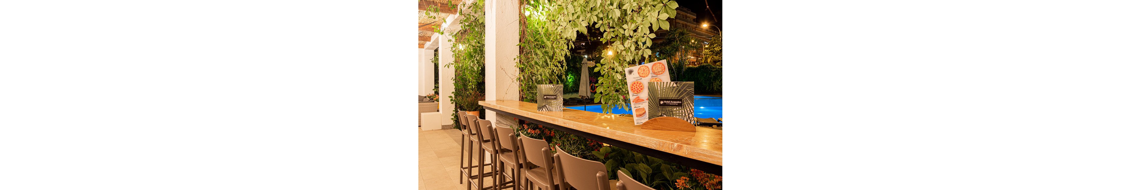An inviting outdoor bar overlooks a brightly lit swimming pool at night. High stools line the wooden counter, which is decorated with lush green plants and colorful flowers. A menu stand with tropical leaf graphics and images of food items sits on the bar surface. Warm ambient lighting and greenery create a relaxed, vacation-like atmosphere in this poolside setting.