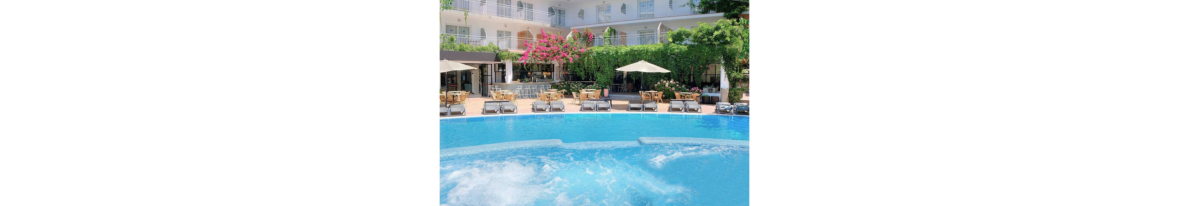 An outdoor swimming pool with bubbling water sits at the center of a bright hotel courtyard. Sun loungers and umbrellas line the poolside terrace, with tables and chairs arranged for outdoor dining. The hotel building rises in the background with balconies and large windows overlooking the scene. Lush green vines and vibrant pink flowers add color and a relaxed, Mediterranean resort atmosphere.
