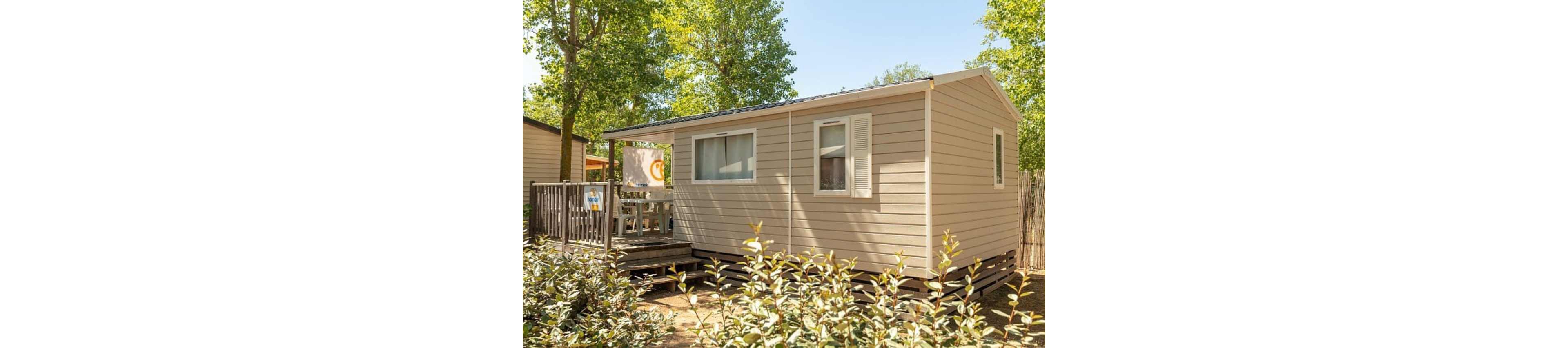 A beige mobile home stands in a sunny outdoor setting surrounded by green trees and shrubs. The structure features a small wooden deck with outdoor furniture partially visible through the railing. Closed shutters and a curtained window give the home a tidy, well-kept appearance. Warm daylight and lush foliage create a relaxed, vacation-like atmosphere.