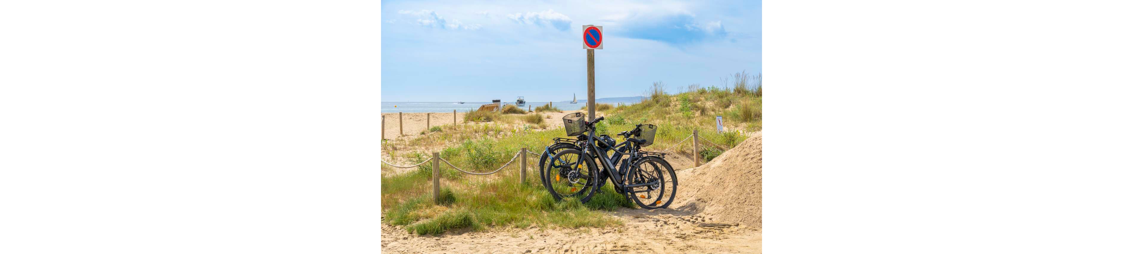Two electric bicycles are parked together near a sandy beach path. A no-parking road sign stands above them, with the sea and a distant sailboat visible in the background. The scene features grassy dunes, wooden posts with rope, and a bright, partly cloudy sky. The overall atmosphere suggests a relaxed coastal outing and eco-friendly transportation.