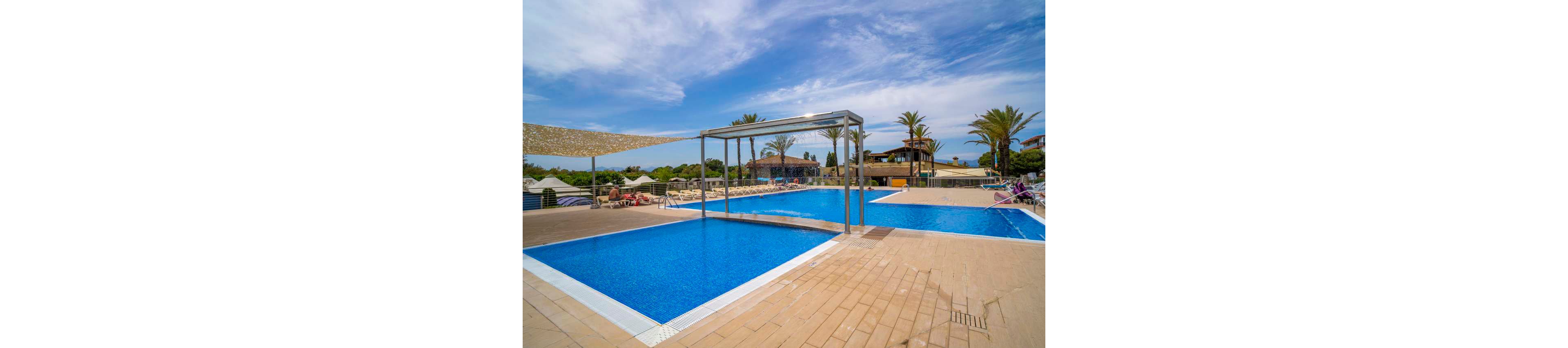 A bright blue resort swimming pool is shown under a clear sky, featuring a modern rectangular metal frame structure over the water. The pool deck is spacious with lounge chairs and umbrellas arranged around the perimeter. Palm trees and low-rise resort buildings are visible in the background, suggesting a warm vacation destination. The overall scene feels calm and inviting, ideal for leisure and relaxation by the water.