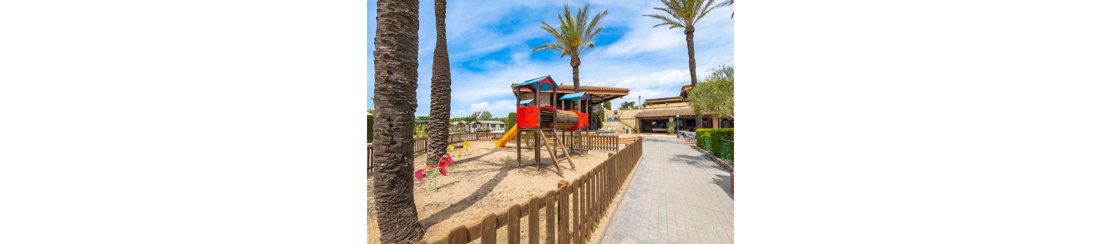 A fenced playground features colorful wooden play structures with slides and climbing elements set in a sandy area. Tall palm trees cast shadows across the sand, creating a warm and inviting atmosphere. A paved walkway runs alongside the playground, leading toward nearby buildings and outdoor seating areas. The bright blue sky with scattered clouds enhances the sunny, relaxed resort-like setting.
