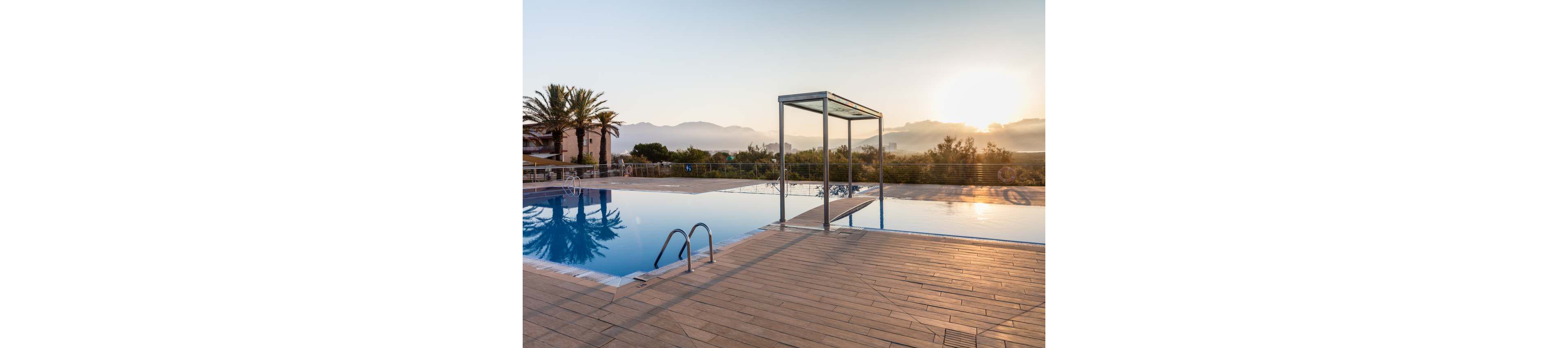 A serene outdoor swimming pool reflects the soft light of sunrise. The scene features a diving board structure and metal pool ladder on a spacious tiled deck. Mountains and trees form a calm natural backdrop, with a few buildings partially visible to the side. The still water and warm sky create a tranquil, resort-like atmosphere.