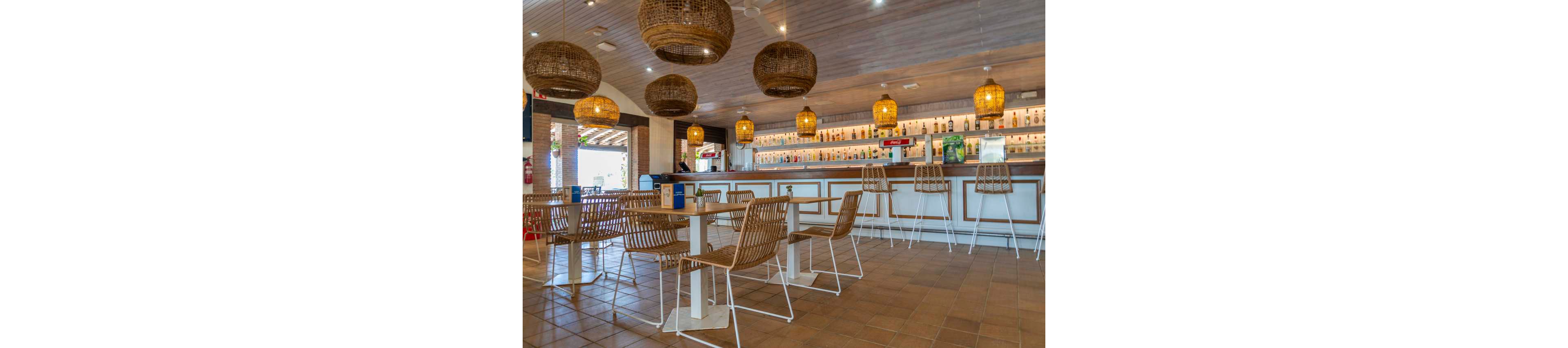A contemporary bar interior features wicker chairs and tables arranged on a tiled floor. The long counter in the background is lined with bottles and illuminated shelving. Large woven pendant lights hang from the wooden ceiling, casting a warm glow over the space. Natural light enters from an open side area, enhancing the relaxed, coastal atmosphere.